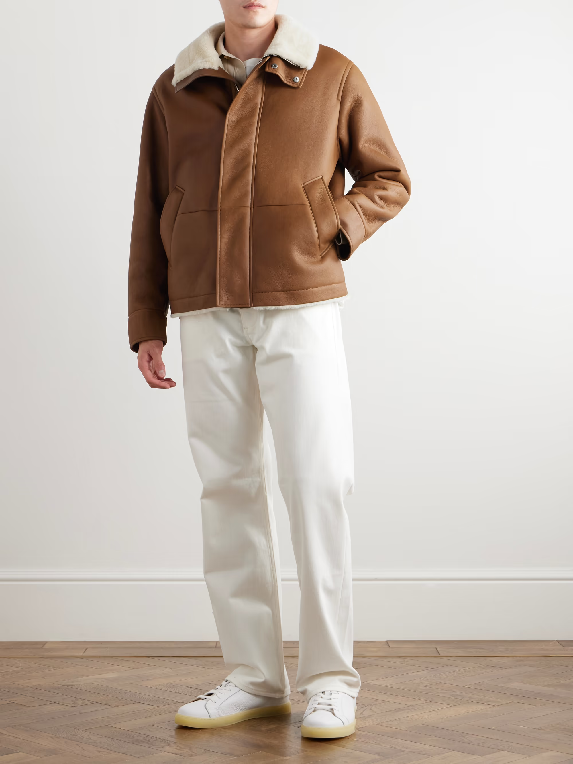 Person wearing a brown leather jacket with a white interior, white pants, and white sneakers on a plain background.