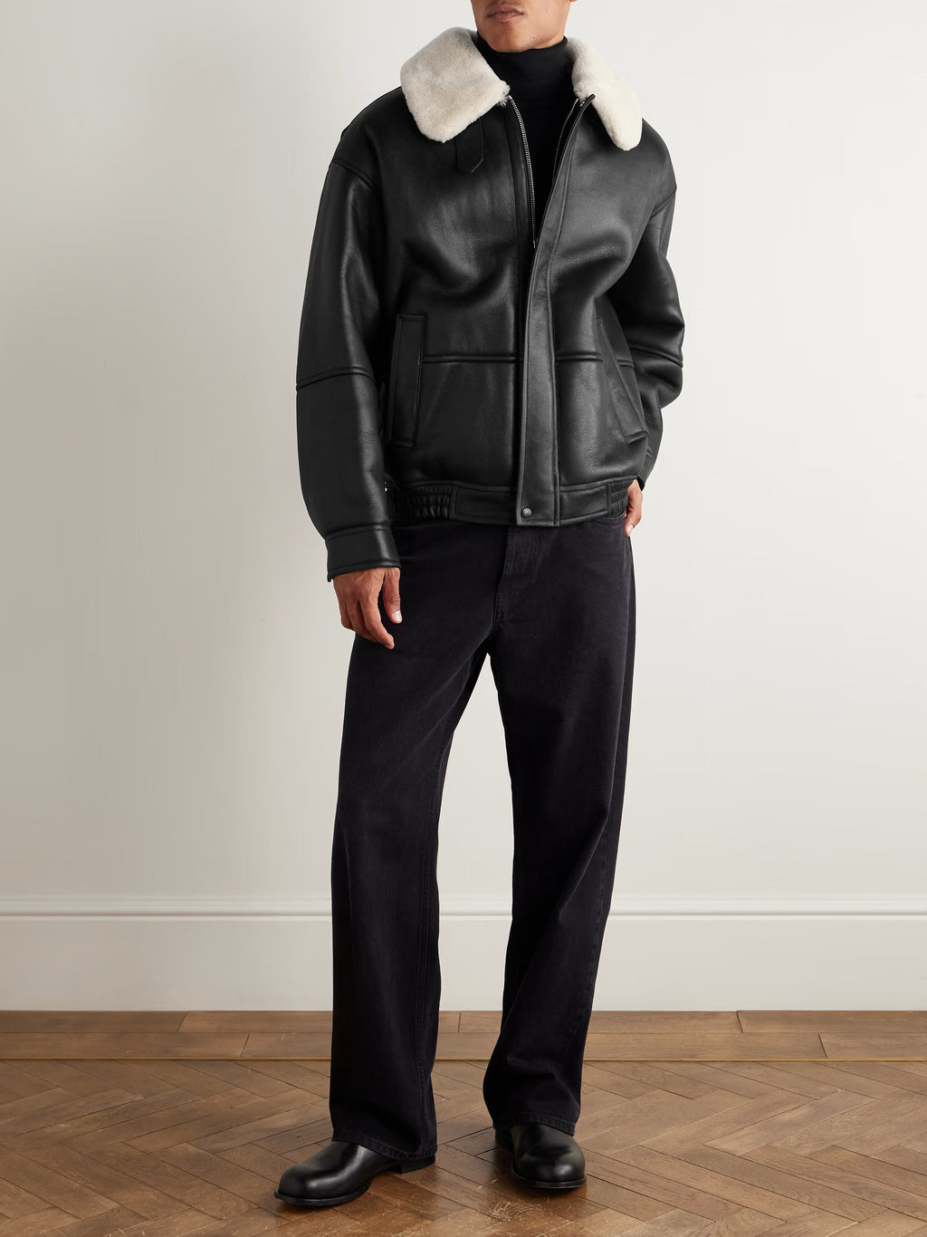 Person wearing a black leather jacket with white fur collar and black pants on a plain background