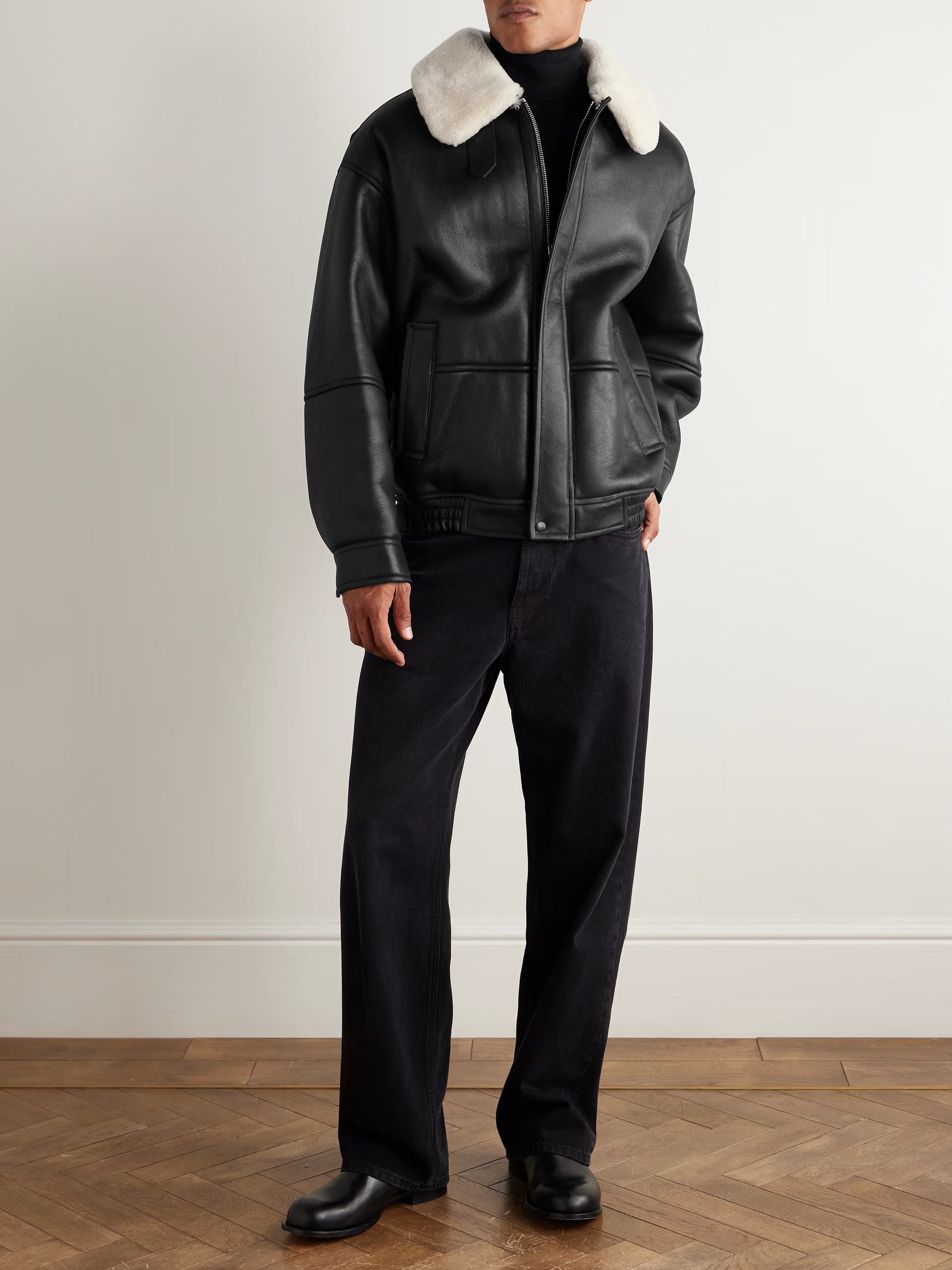 Person wearing a black leather jacket with white fur collar and black pants on a plain background